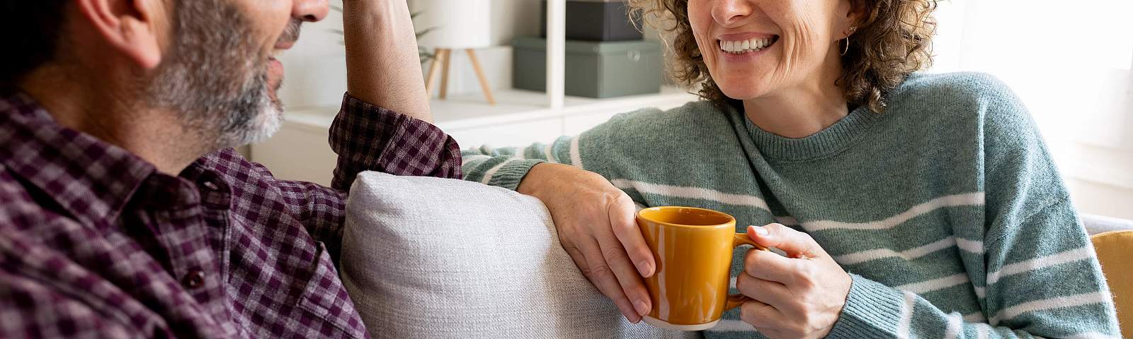 Two people smiling and talking on a couch in a living room setting, one person holding an orange mug