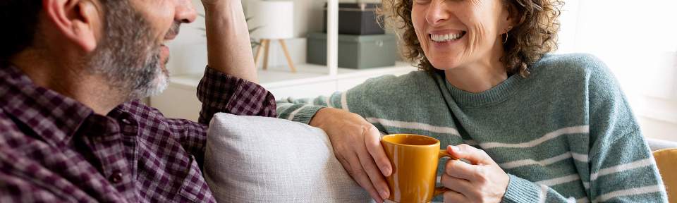 Two people smiling and talking on a couch in a living room setting, one person holding an orange mug